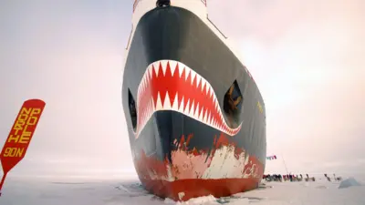 На заднем плане - небо, затянутое облаками. На среднем плане - нос ледокола. На нем нарисована оскаленная пасть. Справа от ледокола стоит толпа людей на льду. Слева - красная табличка, на которой желтыми буквами написано по-английски 